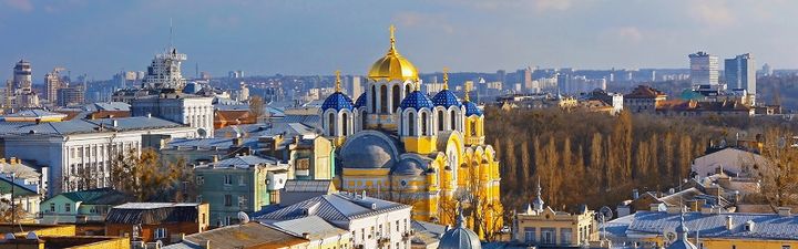 Ein Panoramabild der St. Volodymyr-Kathedrale in Kiew, Ukraine, mit goldenen Kuppeln und blauen Dächern, umgeben von anderen Gebäuden und einer baumbestandenen Fläche, unter einem hellblauen Himmel.
